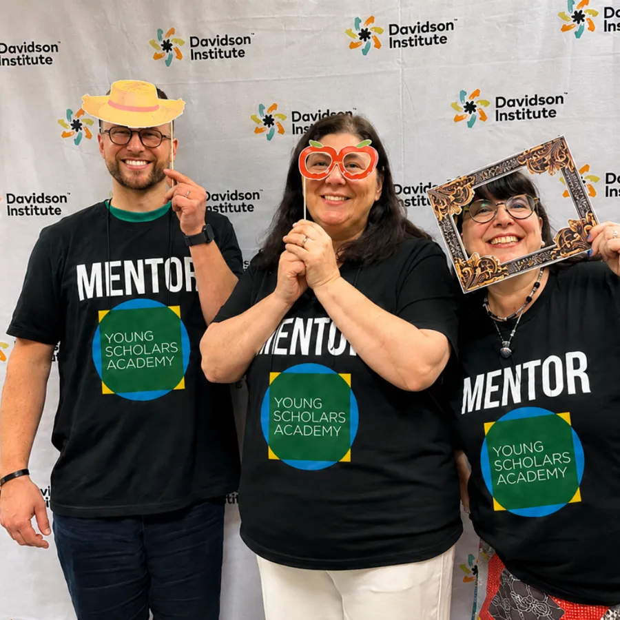 Three Young Scholars Academy mentors in MENTOR shirts posing with photo-booth props at an event